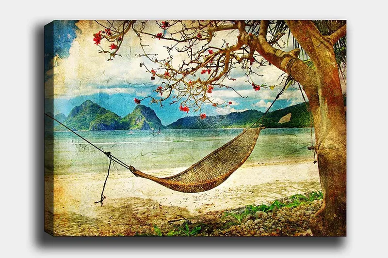 Canvastavla 70x100 cm - Avkopplande hängmatta hängande mellan trädgrenar vid en vacker strand med fjärran berg i bakgrunden, Grön / Brun / Blå